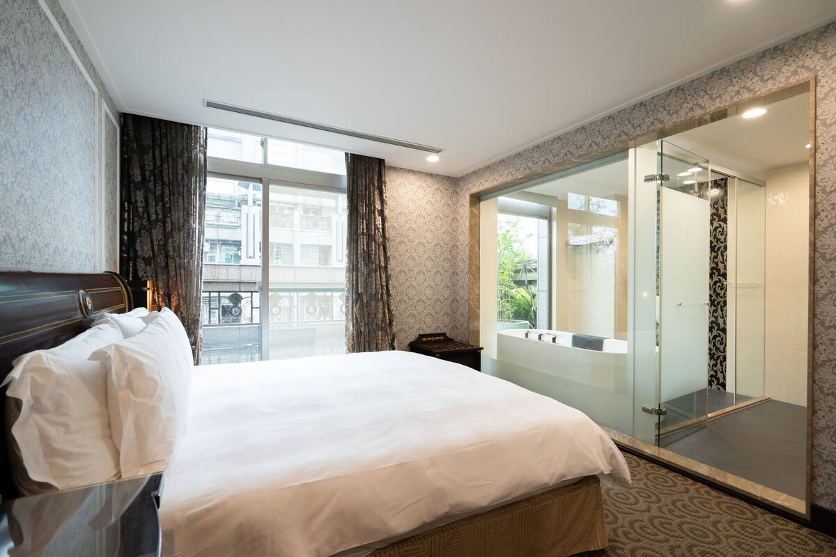 Habitación de hotel con una cama grande, ducha con paredes de cristal y vistas al exterior.