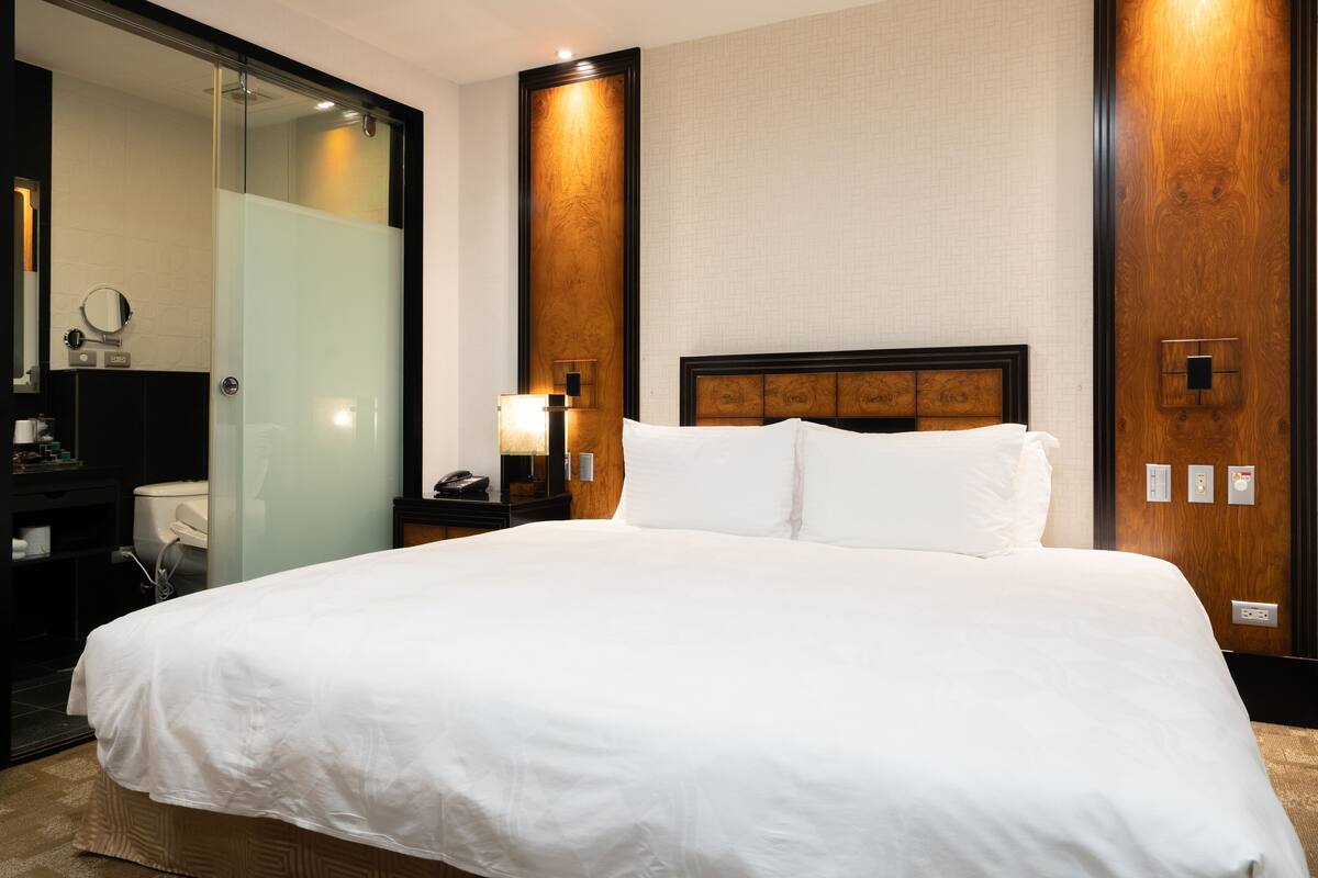 Habitación de hotel con una cama grande, cabecera de madera, mesita de noche con lámpara, espejo y baño con inodoro y lavamanos.