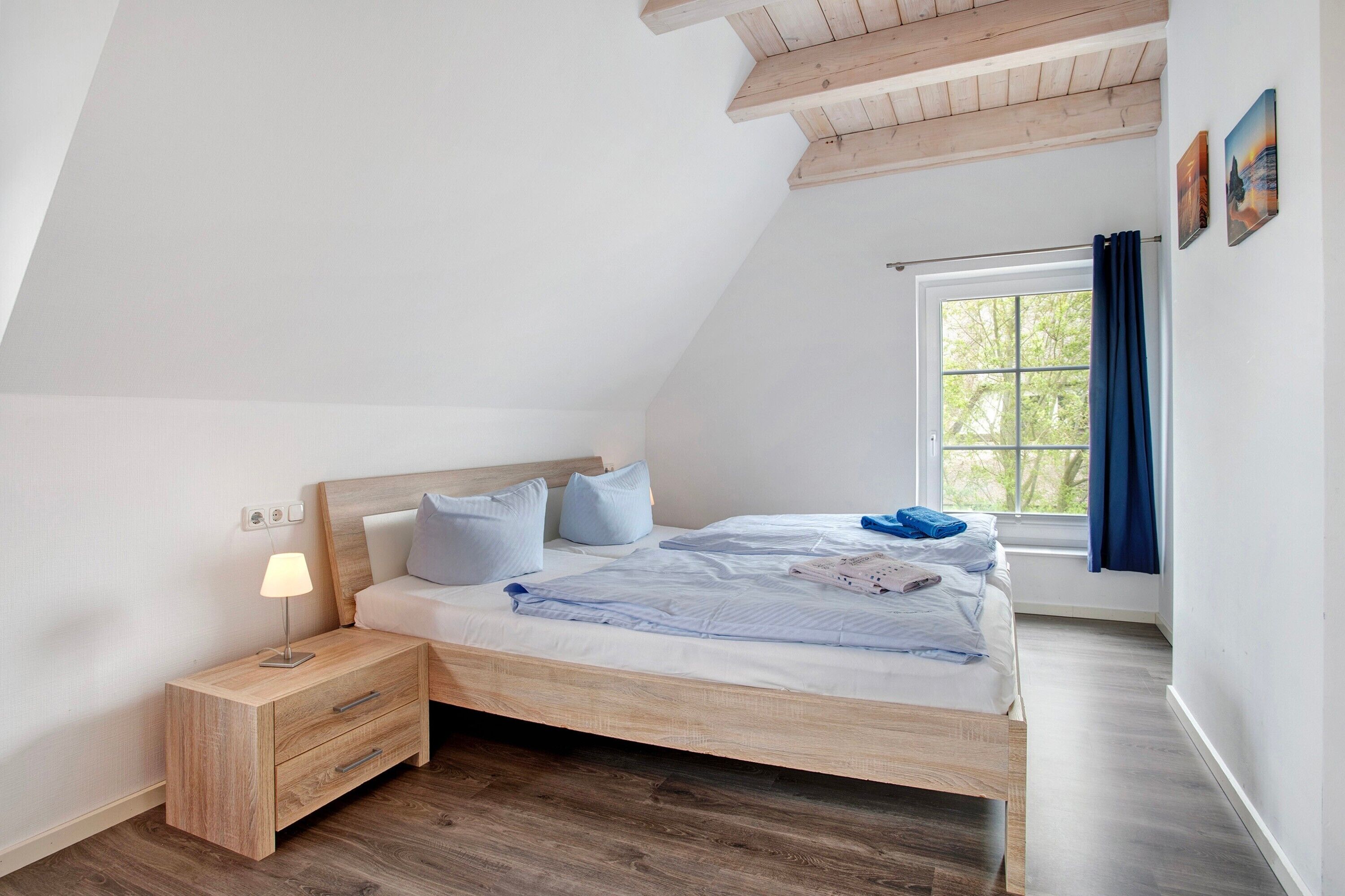 A bedroom with a sloped ceiling, a bed with a wooden frame, a nightstand with a lamp, and a window with curtains.
