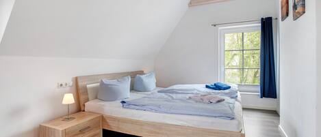 A bedroom with a sloped ceiling, a bed with a wooden frame, a nightstand with a lamp, and a window with curtains.