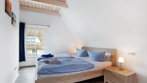 Une chambre avec un plafond en pente, un lit avec une literie bleue, une table de chevet avec une lampe et une fenêtre avec des rideaux.