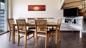 Un coin repas avec une table et des chaises en bois, un tableau représentant un coucher de soleil, un téléviseur posé sur une étagère blanche et un escalier.