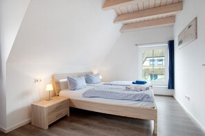 Une chambre avec un plafond en pente, un lit avec une literie blanche, une table de chevet avec une lampe et une fenêtre avec des rideaux.