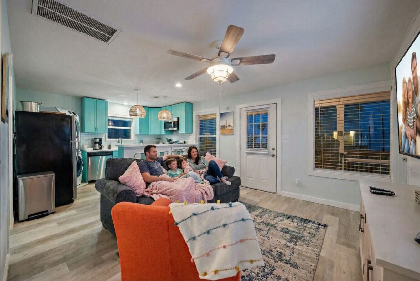 A family in a modern living room with a kitchen in the background, a ceiling fan, and a large window with blinds.