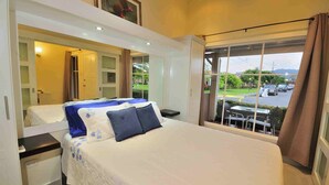 A bedroom with a large bed, a nightstand, a dresser, and a window with a view of the street and greenery outside. - Newly Renovated Condo One Block From Beach (Buenos Aires)