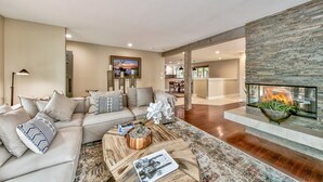 A modern living room with a sectional sofa, a coffee table with magazines, a fireplace with a glass enclosure, and a dining area in the background.