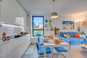A modern kitchen with white cabinets, a dining area with blue chairs, and a blue sofa with orange pillows.