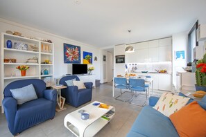 A modern living room with a sofa, armchairs, a coffee table, and a TV. There is a dining area with a table and chairs. The kitchen is visible with cabinets and a hanging light.