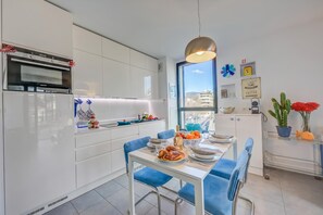 Una cucina moderna con mobili bianchi, forno integrato, tavolo da pranzo con sedie blu e vista attraverso un'ampia finestra.