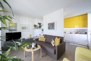 Un salon moderne avec un canapé, une table basse et un téléviseur. Il y a un coin cuisine avec des placards jaunes et un coin repas avec une chaise jaune.