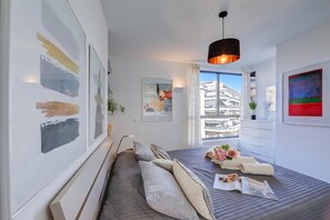 A modern bedroom with a large bed, a bedside table, a hanging light, and a view of the city.