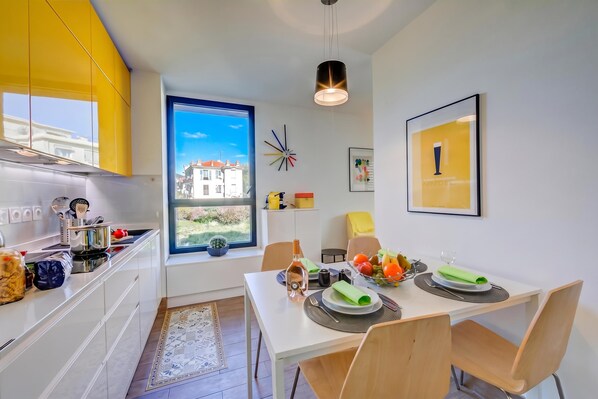 Une cuisine moderne avec des meubles blancs, un coin repas avec une table dressée pour deux personnes et une grande fenêtre donnant sur des immeubles.