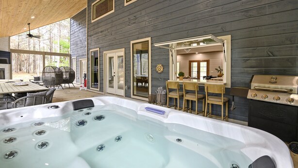 A hot tub in a covered outdoor area with a wooden ceiling and walls, a grill, and a dining area with chairs and a table. - Stunning Modern Cabin (Broken Bow)