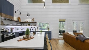 A modern kitchen with a central island, wooden cabinets, and a brown leather couch in the living area. - Stunning Modern Cabin (Broken Bow)