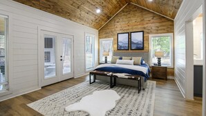A bedroom with a wooden ceiling, a bed with a white and blue bedspread, a white rug, and a white cowhide rug. - Stunning Modern Cabin (Broken Bow)