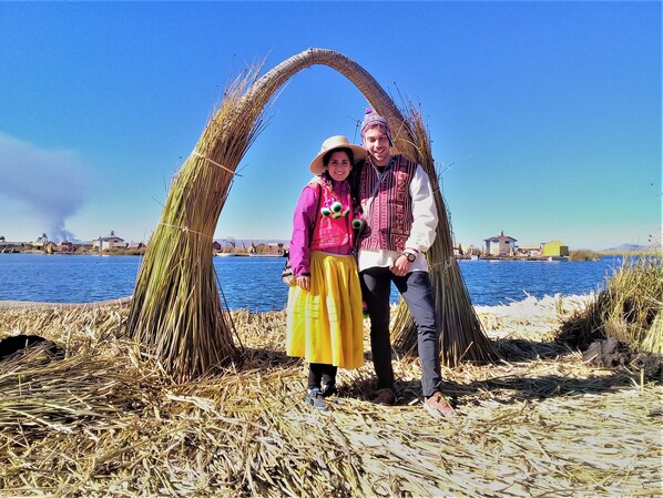 A couple standing in a rural area with a straw archway, a body of water, and buildings in the background.