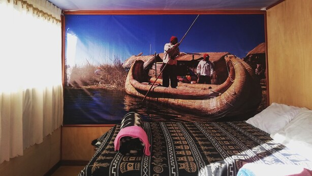 A room with a large mural of a traditional boat scene, a bed with patterned bedding, and a window with sheer curtains.