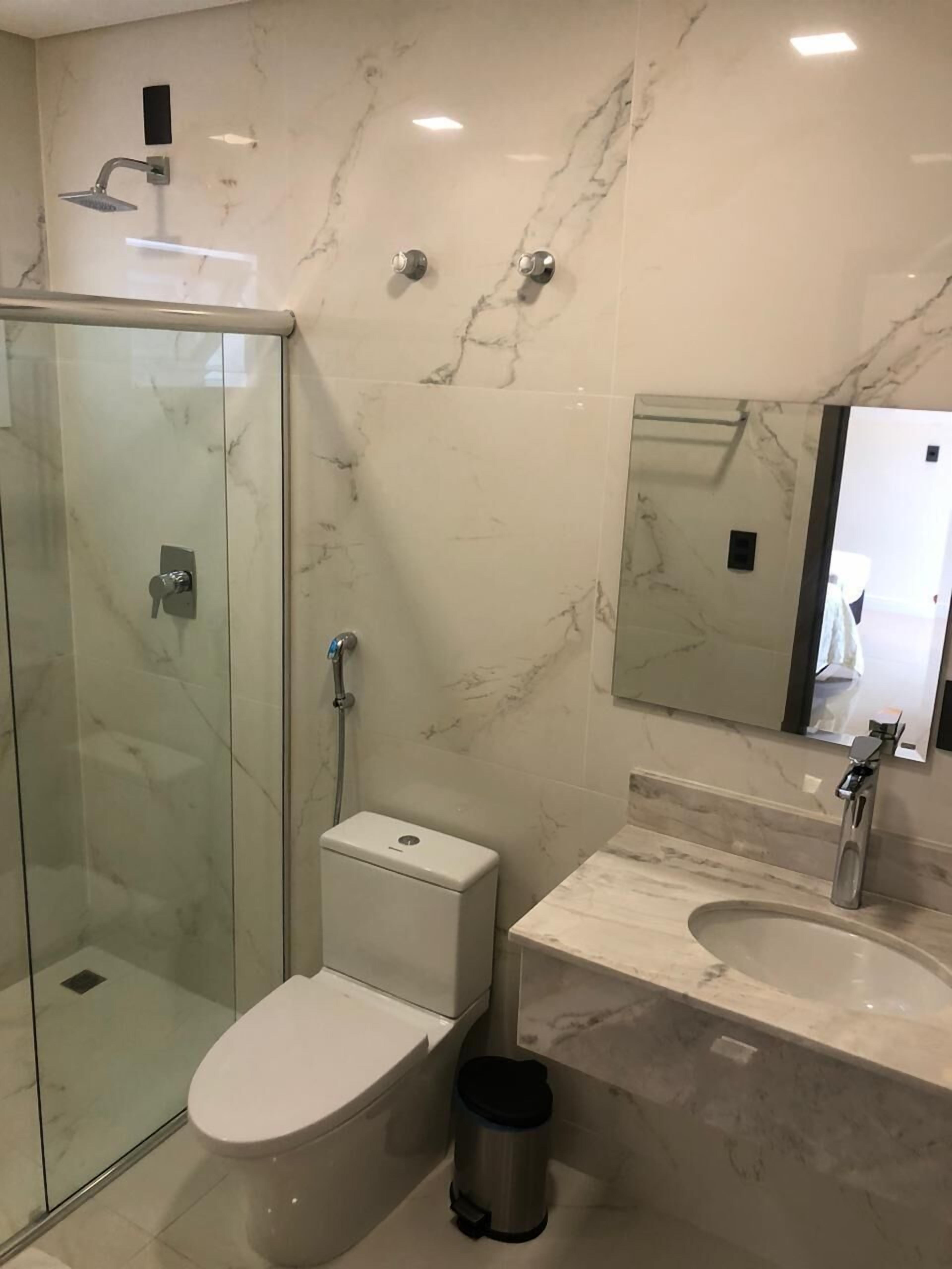 Egy modern fürőszoba, melyben üvegfalas zuhanyzó, WC, bidé és mosdókagyló található, a mosdókagyló felett pedig tükör van.