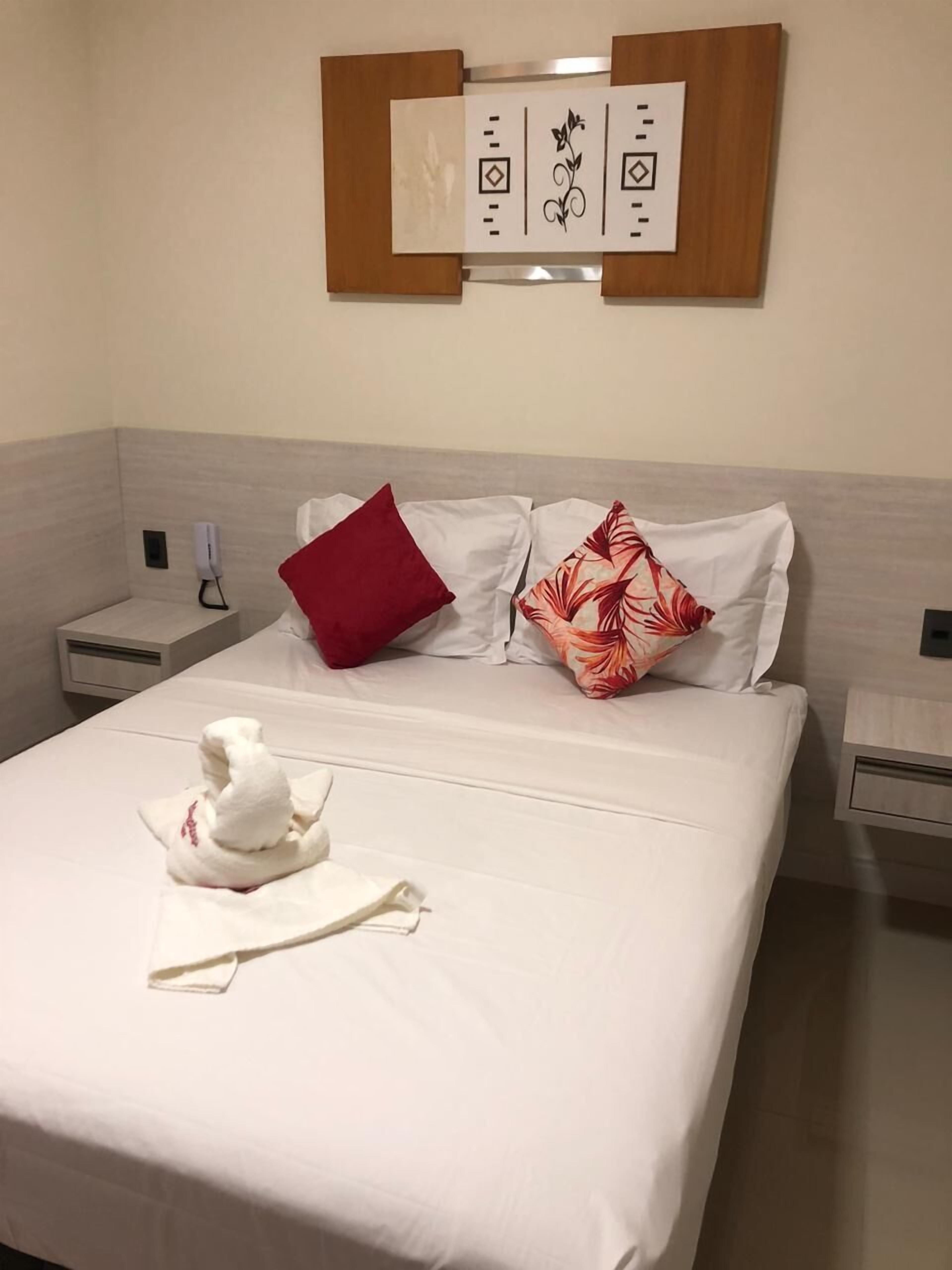 Una habitación de hotel con una cama bien hecha, un cojín rojo y otro estampado con motivos florales, una toalla blanca y una obra de arte colgada en la pared.