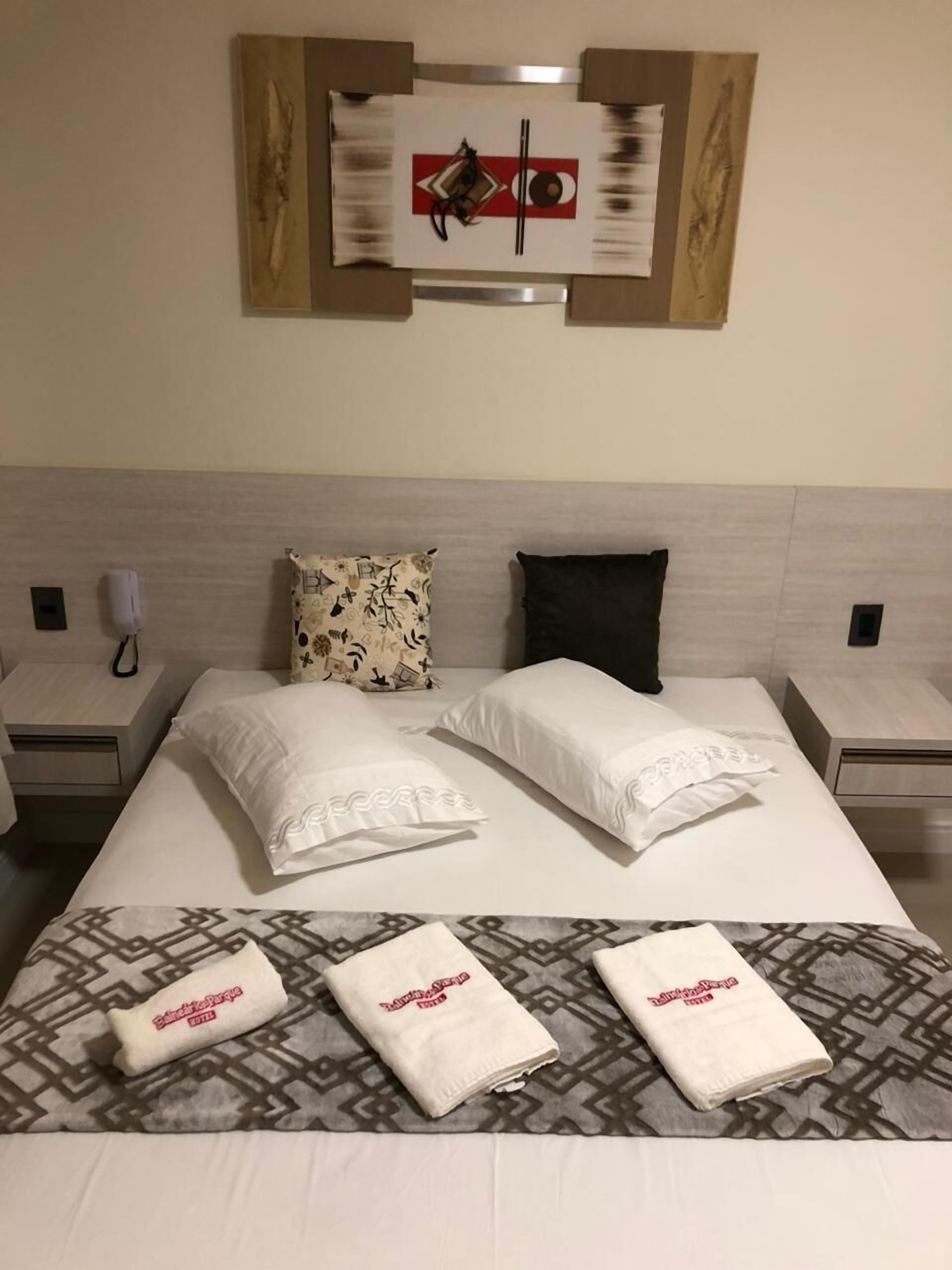 A neatly made bed with two pillows, a patterned throw, and two towels with text. There are two bedside tables with lamps and a wall-mounted artwork above the bed.