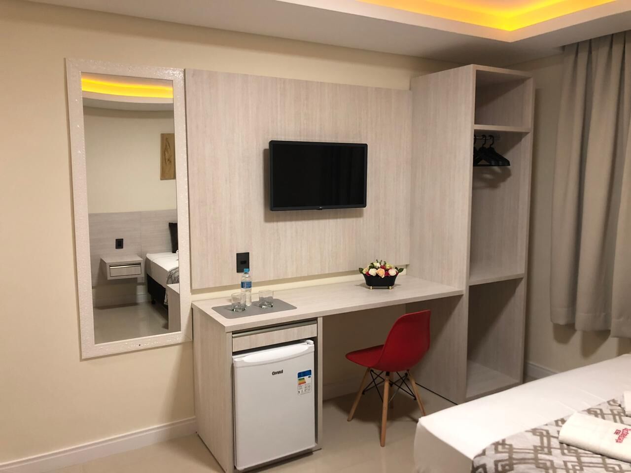 A compact hotel room with a bed, a small desk with a TV, a mini-fridge, a red chair, and a mirror.