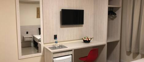 A compact hotel room with a bed, a small desk with a TV, a mini-fridge, a red chair, and a mirror.