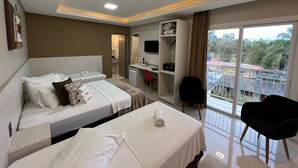 A hotel room with two beds, a desk, a chair, a TV, and a balcony with a view of buildings and trees. - Hotel Balneário do Parque (Penha)