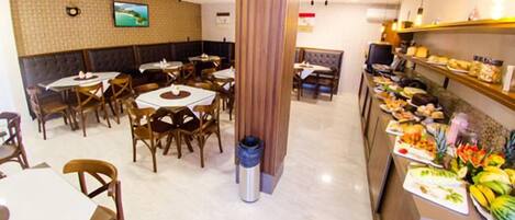 Un restaurante con mesas y sillas redondas, una zona de bufé con diversos alimentos y un televisor montado en la pared.