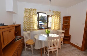 A dining area with a round table, white chairs, a potted plant, and a wooden bookshelf.