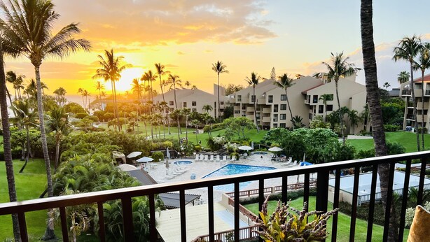 A balcony with a glass table, a vase with flowers, and a view of a pool and resort buildings. - Maui South Kihei New Modern Spacious Ocean View Condo, 5 mins walk to beach! (KIHEI)