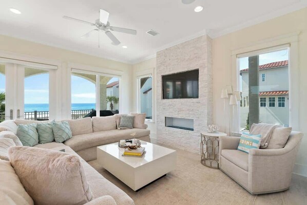 A spacious living room with a large sectional sofa, a coffee table, and a fireplace. There is a ceiling fan, large windows, and a view of the ocean. - Vizcaya 1001 (Santa Rosa Beach)