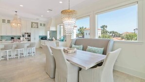 Cocina moderna con armarios blancos, comedor con banco y un amplio ventanal con vistas a palmeras.