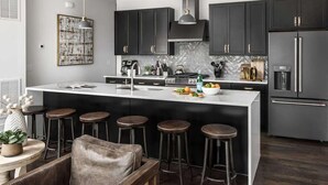 Una cocina moderna con armarios oscuros, una isla grande con taburetes, un refrigerador y una lámpara colgante.
