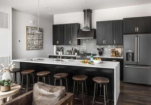 A modern kitchen with dark cabinets, a large island with stools, a refrigerator, and a pendant light. - Tennessee Row - Heart of Gulch - Mins to Broadway (Nashville)