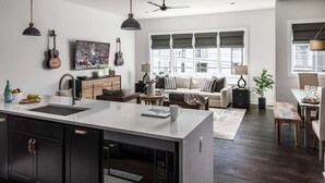 Una cocina moderna con una isla central, una zona de comedor con mesa y sillas, un sofá, y un amplio ventanal con persianas.
