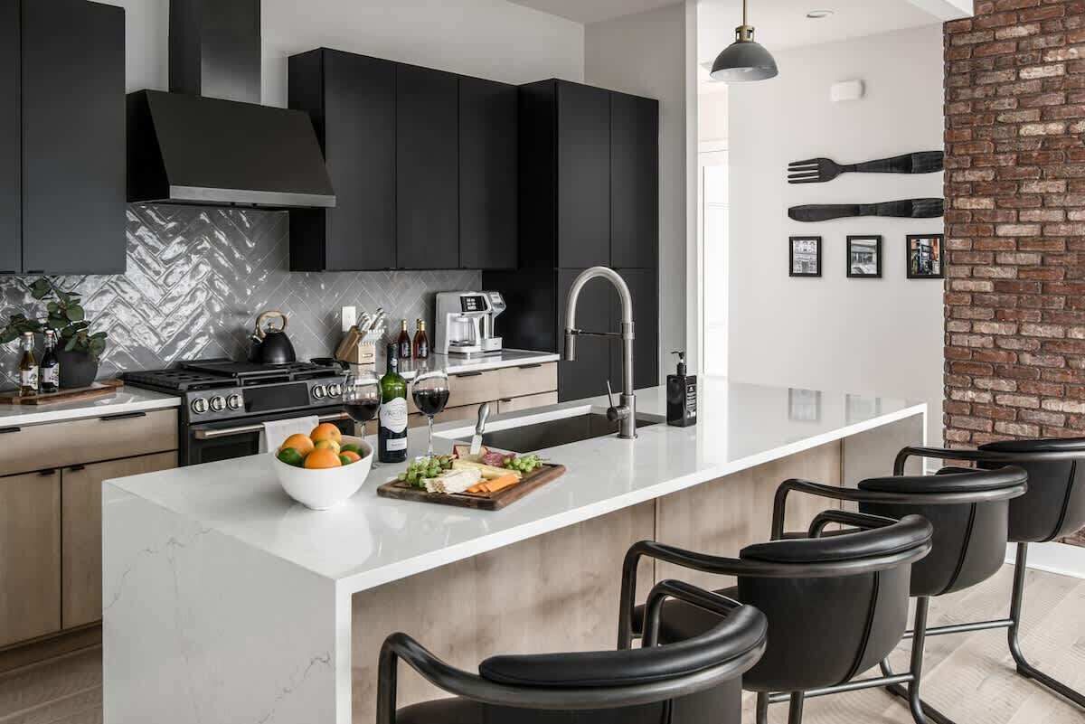 A modern kitchen with dark cabinets, a white island, bar stools, and a brick accent wall.