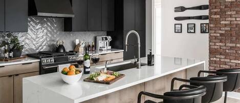A modern kitchen with dark cabinets, a white island, bar stools, and a brick accent wall.
