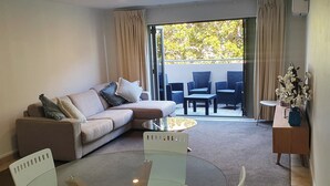 Een moderne woonkamer met een glazen tafel, witte stoelen, een bank met kussens, een salontafel en uitzicht door een schuifpui op een buitenruimte met stoelen en een tafel.