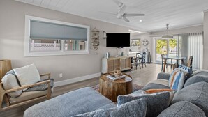 A spacious living room with a large gray sofa, a wooden coffee table, and a TV mounted on the wall. There is a dining area with a table and chairs, and a ceiling fan. - Bungalow by the Beach – Private Pool, Outdoor Lounge, Walk to Sand & Family-Friendly Backyard (Indian Rocks Beach)