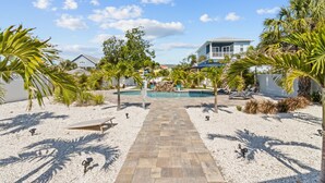 A pathway leading to a pool surrounded by palm trees and a building with a blue roof. - Bungalow by the Beach – Private Pool, Outdoor Lounge, Walk to Sand & Family-Friendly Backyard (Indian Rocks Beach)