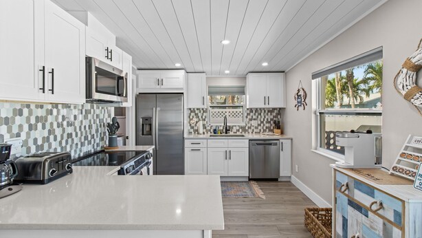A modern kitchen with white cabinets, a central island, stainless steel appliances, and a view of palm trees through the window. - Bungalow by the Beach – Private Pool, Outdoor Lounge, Walk to Sand & Family-Friendly Backyard (Indian Rocks Beach)