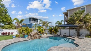 A swimming pool with a stone feature, surrounded by a patio area with lounge chairs and umbrellas, and a two-story house in the background. - Bungalow by the Beach – Private Pool, Outdoor Lounge, Walk to Sand & Family-Friendly Backyard (Indian Rocks Beach)