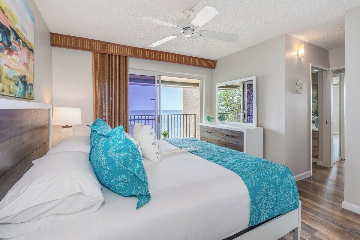 Una habitación de hotel con cama, ventilador de techo, ventana con vistas y puerta que da a otra habitación.