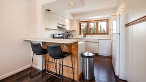 Een moderne keuken met een houten bar, twee zwarte barkrukken, een roestvrijstalen prullenbak en een raam met uitzicht op bomen.