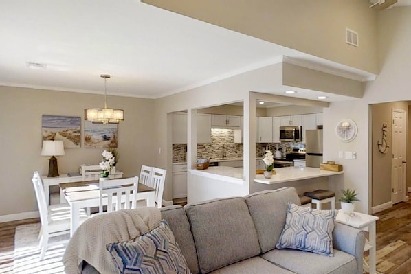 A spacious living room with a gray sofa, a dining area with white chairs, and a kitchen with white cabinets and a stone backsplash. - Best Location-Shipyard, Walk to Beach, Updated, Private Deck, Pool, Golf & Tennis, Guest Favorite (Hilton Head Island)
