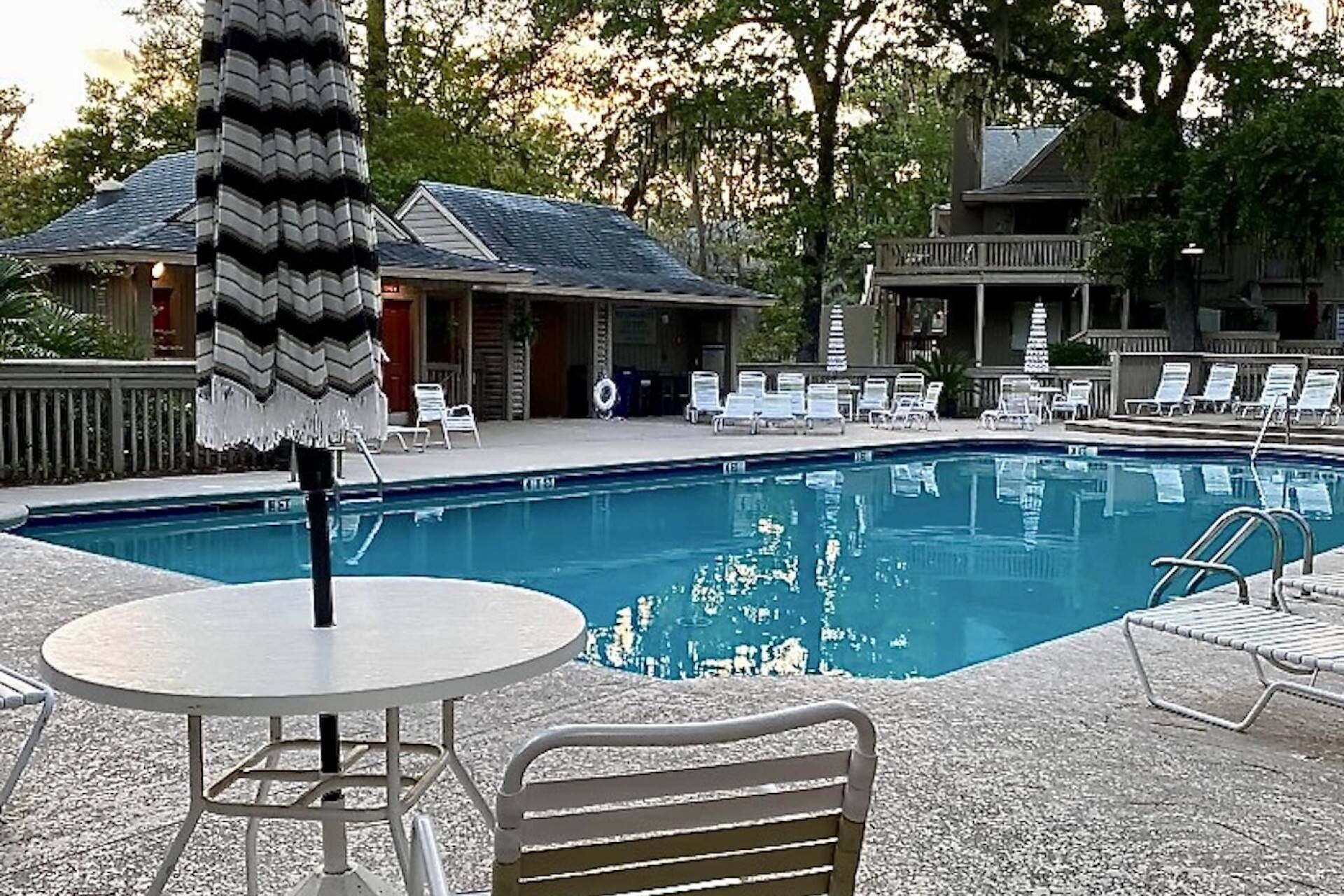 Una piscina con sillas de descanso y una mesa, rodeada de árboles y edificios.