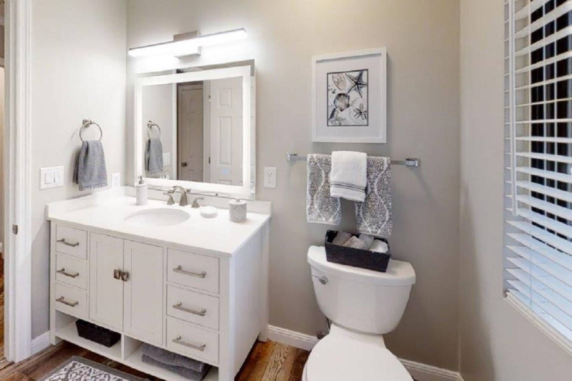 Une salle de bain avec un meuble-lavabo blanc, un grand miroir, des toilettes et une œuvre d’art encadrée accrochée au mur.