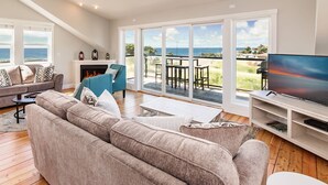 A spacious living room with a large sofa, a television, and a fireplace. There is a balcony with a view of the sea.