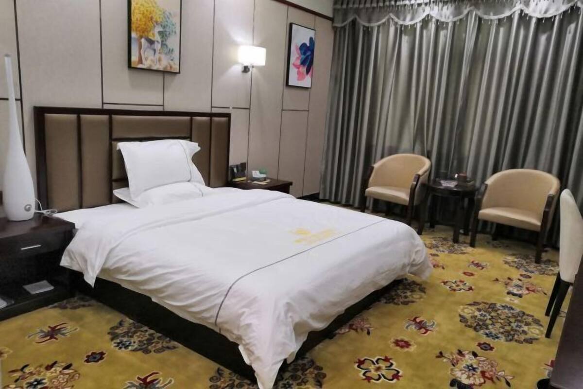 A hotel room with a bed, a nightstand, a lamp, a chair, a small table, and a curtain.
