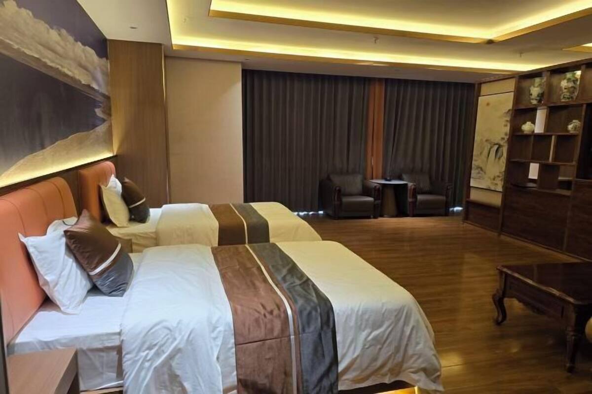 A hotel room with two beds, a small table, a chair, and a bookshelf.